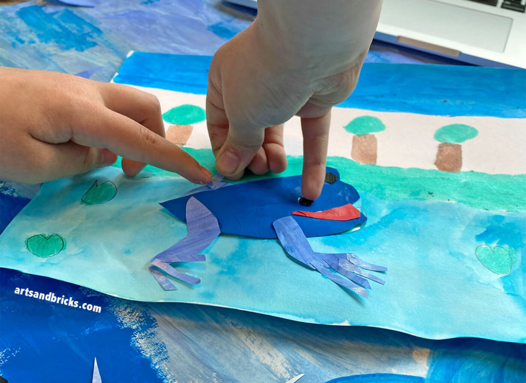 Painted with watercolor paint, the trees, sky, and pond are in the background of this collage project for kids. The frog is in the foreground, and glued layers of pre-painted cut paper form the frog's shape.