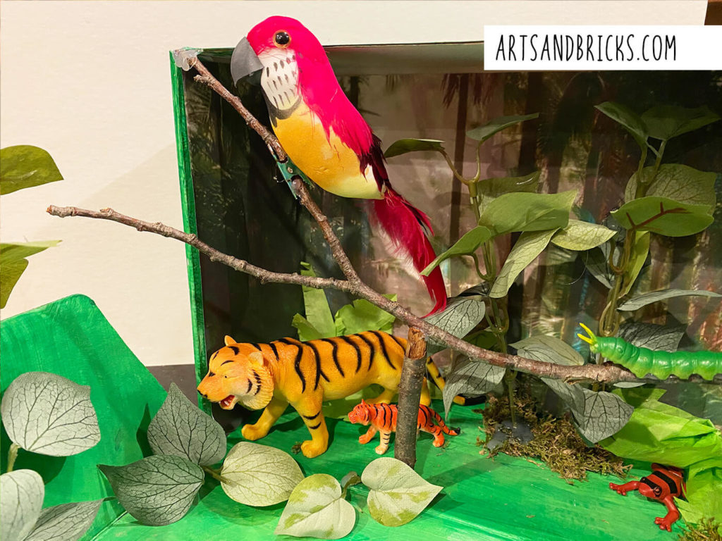 Learn how we made this Jungle Diorama for school using branches, leaves, tissue paper, plastic animals, mosaic, paint and a printed jungle background.